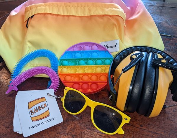 Photo of several items arranged in front of a small soft backpack: there are sunglasses, ear defenders, some colourful fiddle toys, and set of communication cards with the one on top reading 'I want a snack' under a picture of a yellow packet.